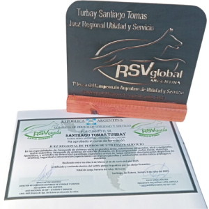 Fotografía que muestra una placa de reconocimiento y un diploma de egresado otorgados a Santiago Tomás Turbay por la organización RSV Global Argentina. La placa tiene forma rectangular, con base de madera, y lleva grabado en relieve el nombre del instructor, su título como Juez Regional de Perros de Utilidad y Servicio, y su participación en la Primera Fecha del Campeonato Argentino de Utilidad y Servicio, realizada en Santiago del Estero los días 4, 5 y 6 de julio de 2025.

Debajo, se exhibe el diploma oficial impreso en papel, con logotipos de RSV Global Latin America y Argentina, donde se certifica la aprobación del curso teórico-práctico con contenidos sobre búsqueda de víctimas, detección de sustancias, y trabajo con unidades de seguridad e intervención.